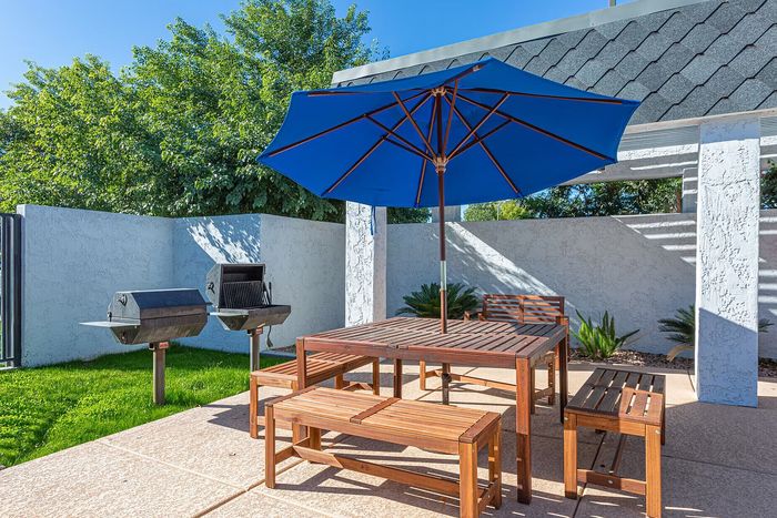 Inviting outdoor patio with a wooden dining set and vibrant blue umbrella beside twin grills, perfect for entertaining and grilling amidst lush greenery.
