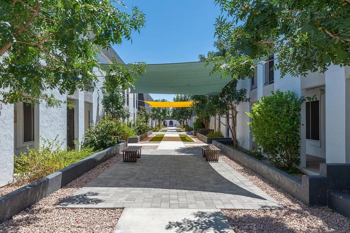 Charming outdoor walkway with lush greenery and shaded seating areas, nestled between modern apartments in serene environment.