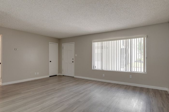 Bright and spacious living room with elegant wooden flooring, large window with vertical blinds, and neutral-toned walls.