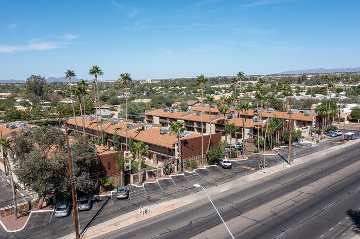 Aerial view of stylish apartment complex with terracotta roofs and lush palm trees, set against a scenic landscape under a clear blue sky.