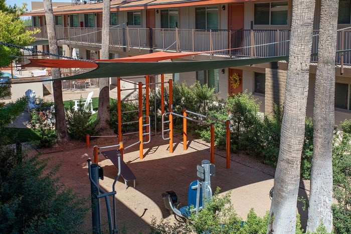 Inviting outdoor fitness area with modern exercise equipment, shaded by colorful canopies, surrounded by lush greenery and cozy apartments.