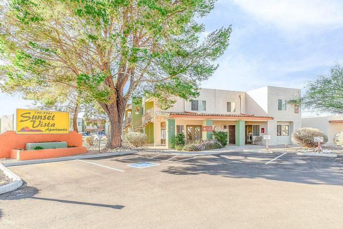 Inviting entrance of Sunset Vista Apartments with vibrant landscaping and ample parking under a clear blue sky.