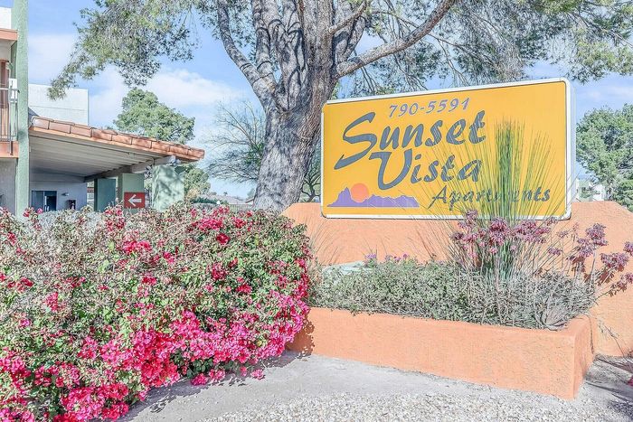 Charming entrance to Sunset Vista Apartments with vibrant flowers and appealing signage under a picturesque tree.