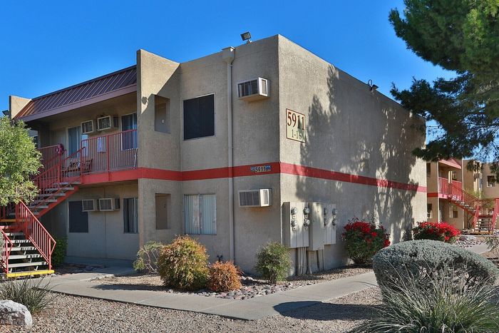 Charming two-story apartment building with stucco exterior, red staircases, and lush landscape featuring vibrant shrubs and trees.