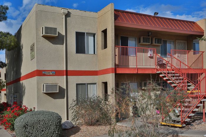 Charming two-story apartment building with vibrant red accents, surrounded by lush greenery and flowers, offering a cozy and inviting atmosphere.