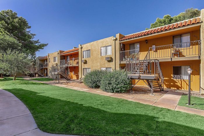 Charming apartment complex with unique orange and beige exterior, featuring spacious balconies, lush green landscaping, and a clear blue sky.