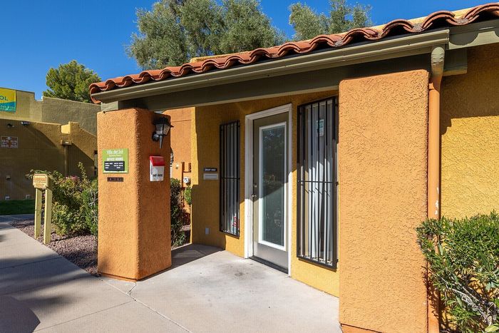 Charming entrance to Villa Del Sol with vibrant orange stucco walls, surrounded by lush greenery and clear blue skies.