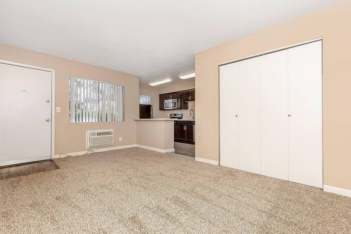 Inviting living space with plush carpet, neutral walls, and an adjacent modern kitchen featuring dark wood cabinetry and stainless steel appliances.