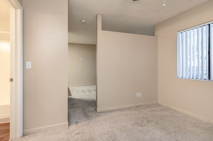 Bright and spacious apartment interior featuring cozy carpeting, a ceiling fan, and natural light streaming through vertical blinds.