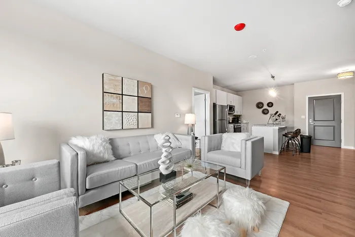 Modern and inviting living room with stylish gray furniture, a chic glass coffee table, and a cozy open-plan kitchen in a bright and airy apartment.