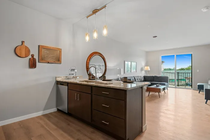 Modern apartment interior featuring a sleek kitchen island with pendant lighting, overlooking a cozy living area with a lush balcony view.