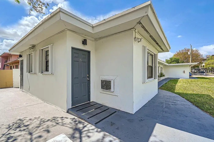 Charming white bungalow with modern gray door, surrounded by lush green lawn and blue skies.