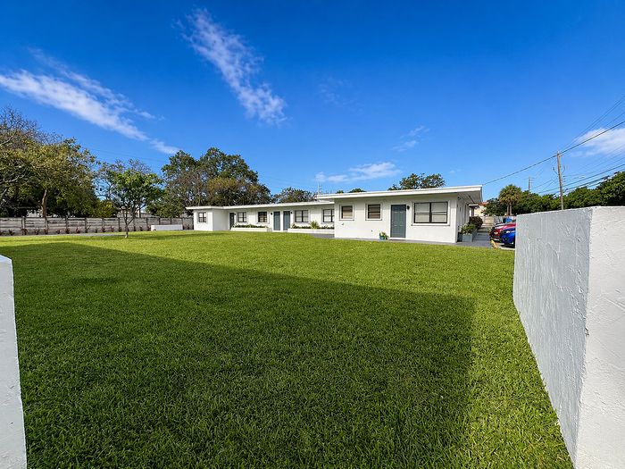 Charming single-story white apartment with a spacious, lush green lawn under a clear blue sky, perfect for outdoor relaxation.