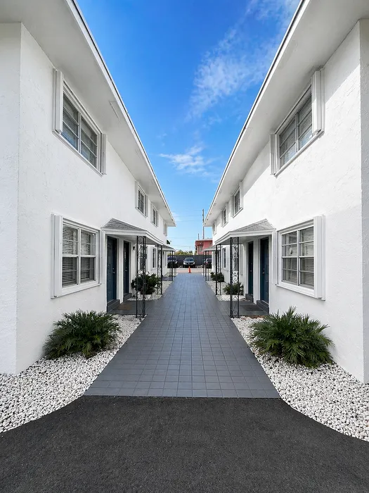 Charming apartment entrance with clean white walls, tiled pathway, and neatly landscaped plants under a clear blue sky.