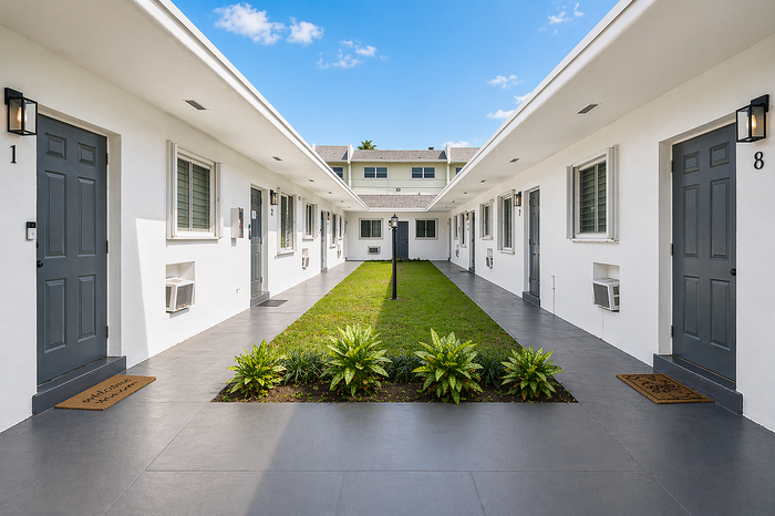 Charming courtyard surrounded by modern apartments with sleek grey doors, lush greenery, and welcoming ambiance under a bright blue sky.