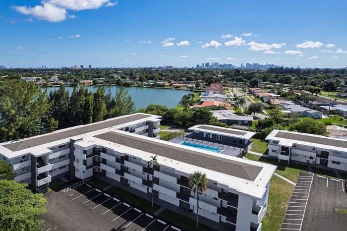 Charming apartment complex with a central pool, nestled by a serene lake and lush greenery, offering picturesque views of the distant city skyline.