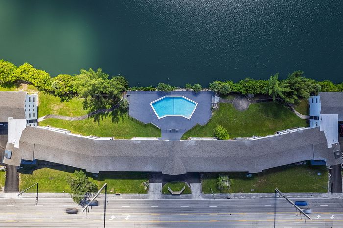 Aerial view of a lakeside apartment complex with a stylish geometric pool, lush greenery, and scenic waterfront setting.