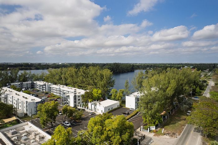 Scenic aerial view of modern apartment complex surrounded by lush greenery, with a tranquil lake under a vibrant blue sky.