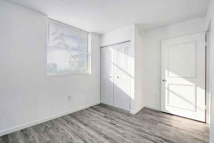 Bright empty room with sleek wood flooring, large window with blinds, and white double-door closet; perfect for a cozy bedroom design.