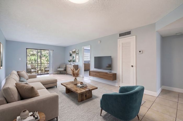 Elegant living room with chic decor, a cozy beige sectional, stylish teal armchair, and a serene view of lush greenery through sliding glass doors.