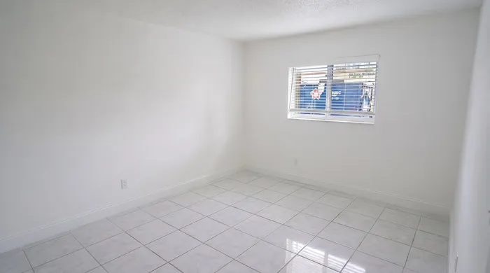 Bright, tiled room with clean white walls and a large window, offering ample sunlight and versatile space.