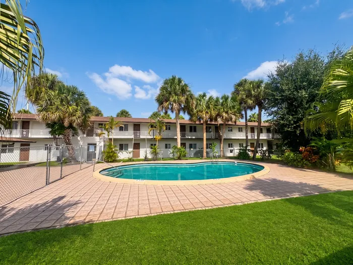 Charming apartment complex with inviting outdoor pool surrounded by lush palm trees and vibrant greenery, perfect for relaxing in the sun.