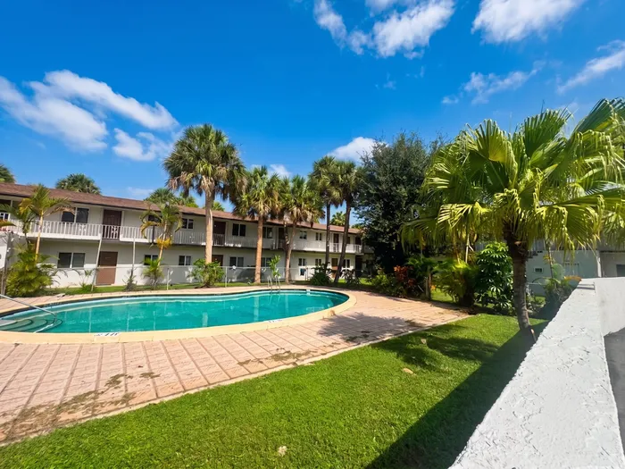Charming apartment complex with a sparkling pool, surrounded by lush palms and vibrant greenery, under a bright blue sky.