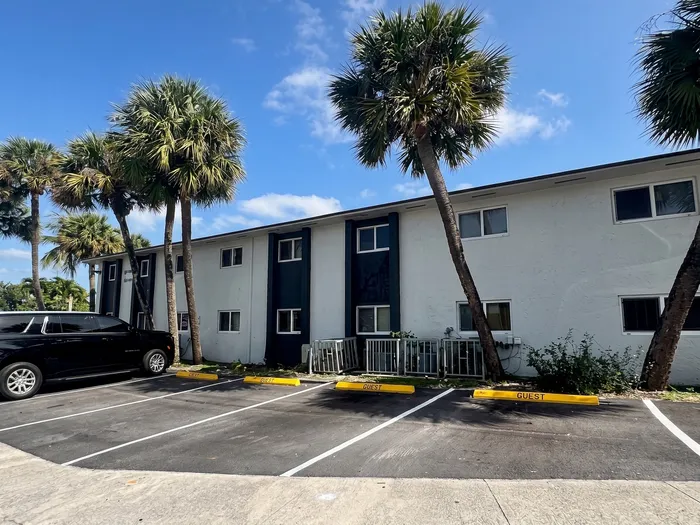 Charming apartment building surrounded by lush palm trees with convenient guest parking and sunny skies.
