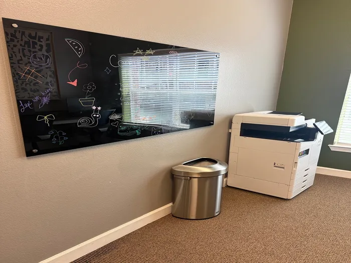 Chic corner office space featuring a sleek blackboard with colorful doodles, a modern printer, and a stainless steel trash can for a productive environment.