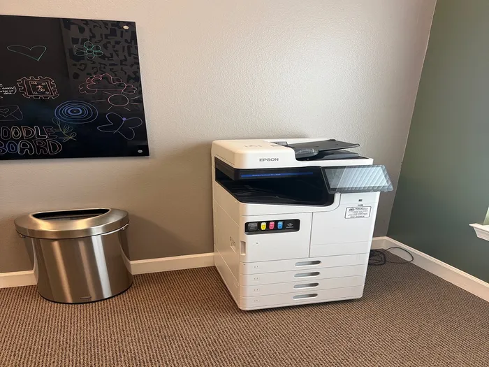 Modern office nook with a sleek Epson printer beside a stylish waste bin and creative doodle board on the wall.