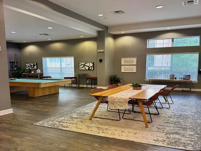 Chic and spacious lounge area featuring a sleek wooden dining table, modern art pieces, and a stylish pool table, bathed in natural light.