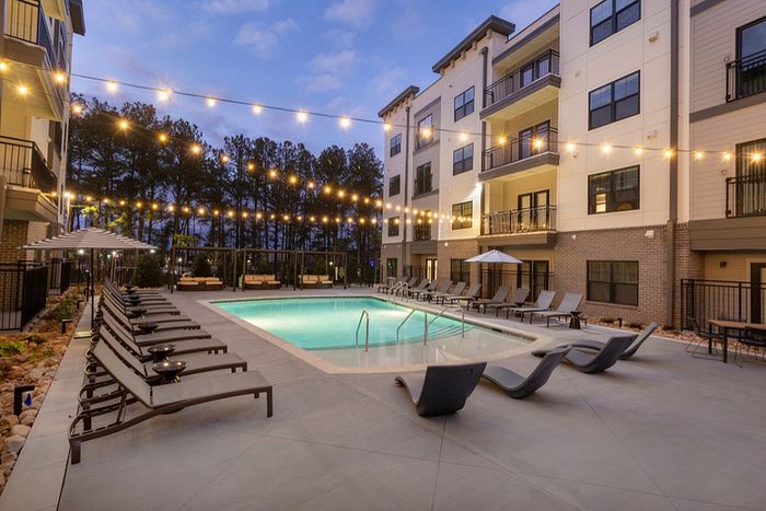 Elegant apartment complex with twinkling patio lights illuminating a serene swimming pool surrounded by stylish lounge chairs and umbrellas at dusk.