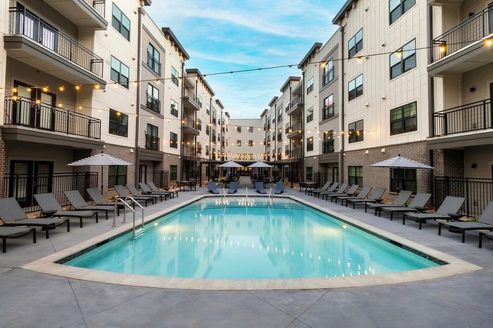 Luxurious apartment complex with a sparkling pool, surrounded by modern balconies and elegant lounge seating under stylish umbrellas.