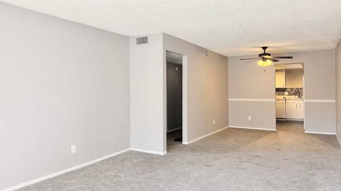 Inviting living space with stylish ceiling fan, cozy carpet, and a glimpse into a modern kitchen featuring sleek cabinetry and backsplash.