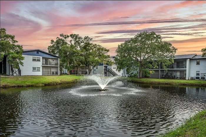 Picturesque apartment complex with serene pond and fountain, surrounded by lush greenery under a stunning sunset sky.