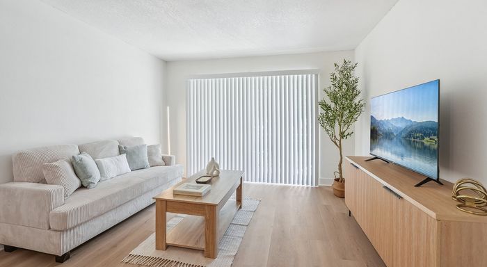 Bright and inviting living room featuring a cozy beige sofa, stylish wooden coffee table, large flat-screen TV, and natural light streaming through vertical blinds.