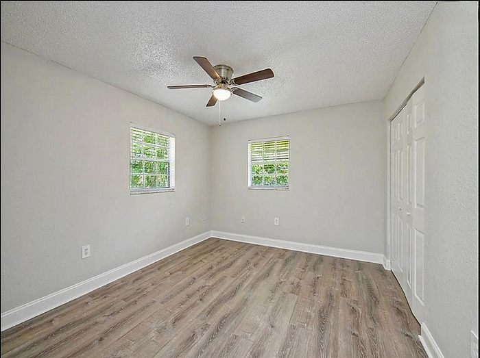 Bright and airy bedroom with sleek wood flooring, ceiling fan, and natural light pouring through two windows.