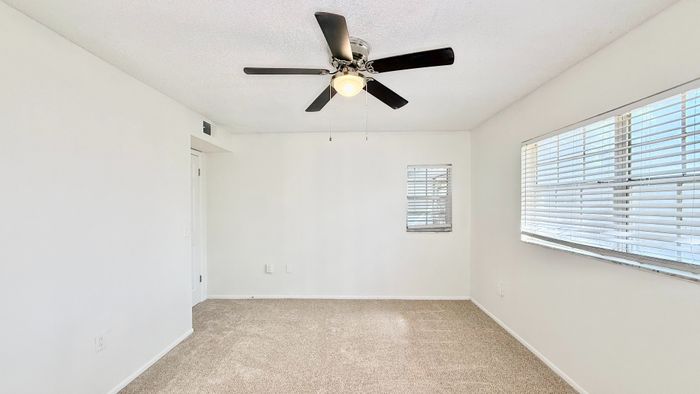 Bright, open room with soft carpet, white walls, and a modern ceiling fan; ample natural light streaming through large windows.