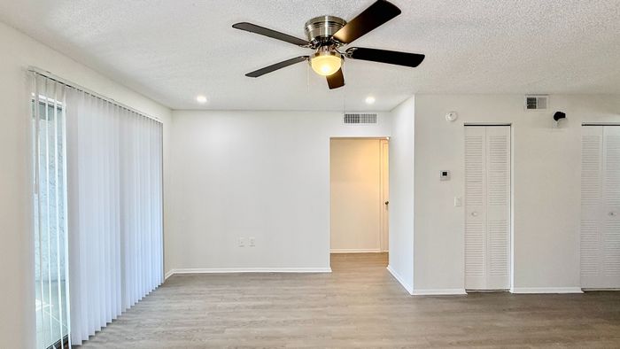 Spacious living room featuring elegant wood flooring, a modern ceiling fan, and large sliding glass doors with vertical blinds for abundant natural light.