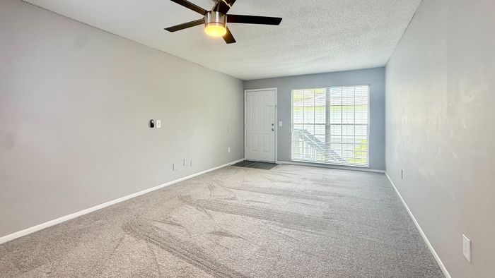 Bright and inviting living room with modern ceiling fan, plush carpet, and large window providing ample natural light.