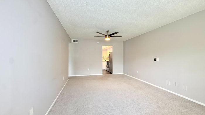 Spacious living room with contemporary ceiling fan and plush carpeting, leading into a modern kitchen with stainless steel appliances.