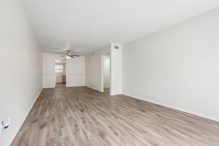 Spacious living area featuring elegant plank flooring, neutral walls, and a ceiling fan with light, leading into a bright kitchen.