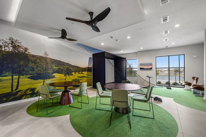 Chic apartment communal area with round tables, green seating, and panoramic nature mural, featuring a golf simulator and large windows offering serene views.