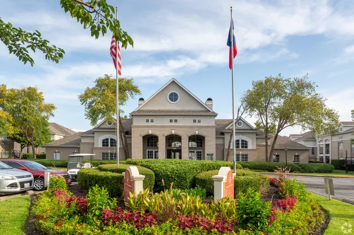 Inviting residential complex entrance with manicured gardens, elegant architecture, and vibrant flags waving under a clear blue sky.