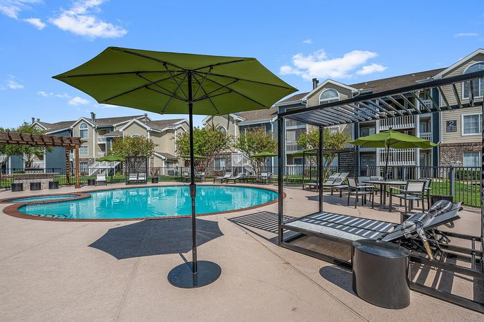 Inviting apartment complex with a sparkling outdoor pool, sun loungers, green umbrellas, and shaded seating areas, perfect for relaxation and entertainment.