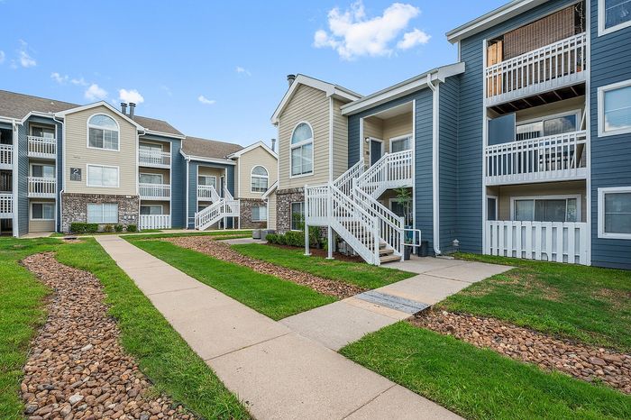 Charming apartment complex with modern architecture, featuring inviting balconies, well-maintained lawns, and a serene pathway under a clear blue sky.