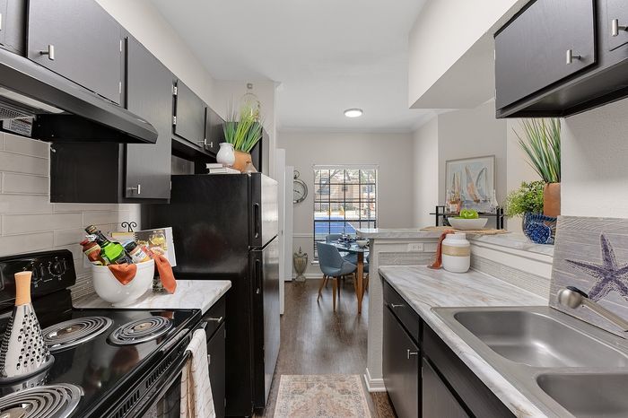Modern kitchen with sleek black cabinets and appliances, leading to a sunlit dining area featuring stylish d&eacute;cor and large window views.