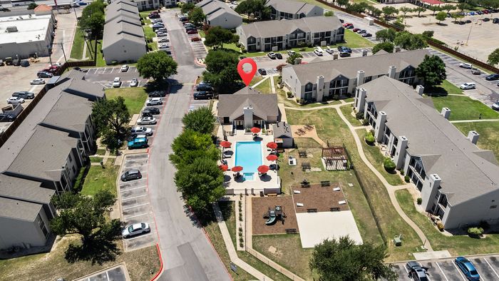 Aerial view of a vibrant apartment complex featuring a central swimming pool with red umbrellas, surrounded by modern two-story buildings, ample parking, and lush green spaces.