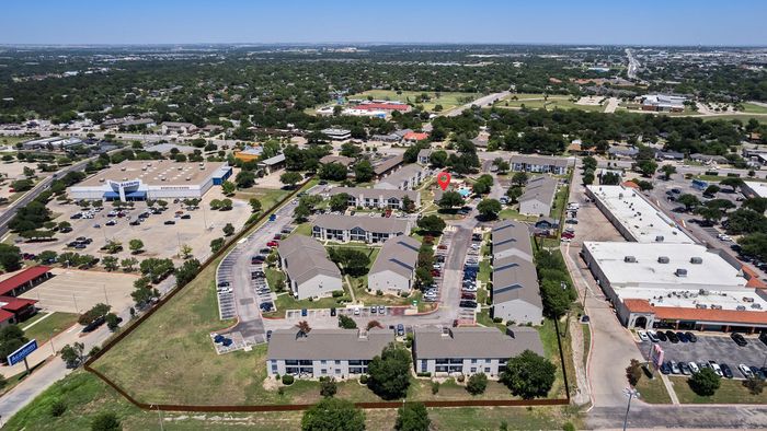 Aerial view of a vibrant apartment complex with a central swimming pool, surrounded by lush greenery and ample parking, conveniently located near shopping centers.