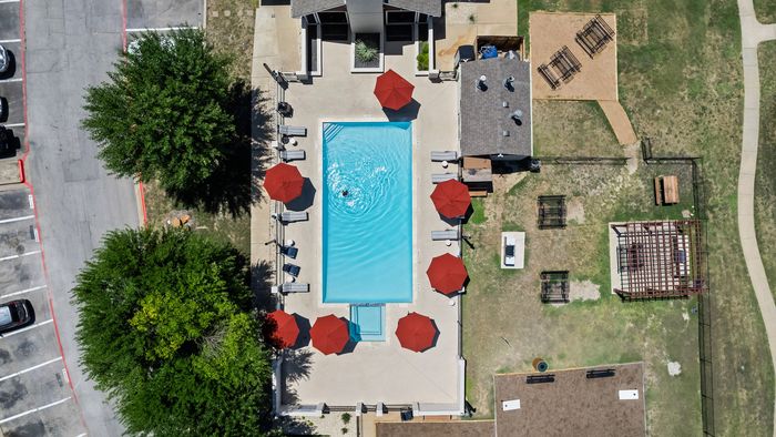 Aerial view of a luxurious apartment complex pool area with inviting lounge chairs, vibrant red umbrellas, and lush greenery, offering a perfect oasis for relaxation and leisure.